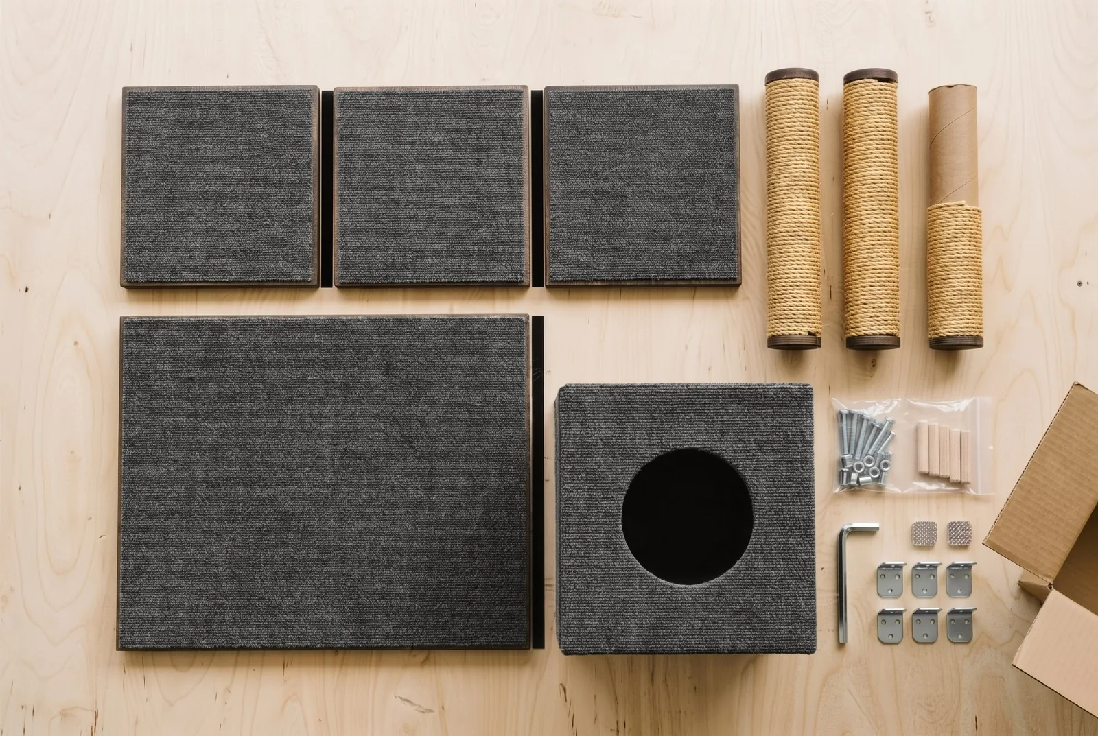 The parts of a Ridgeline laid out on a plywood table: three carpeted platforms, a carpeted base, a hideaway cube, three sisal posts, and a bag of bolts and dowels with brackets and an Allen key.